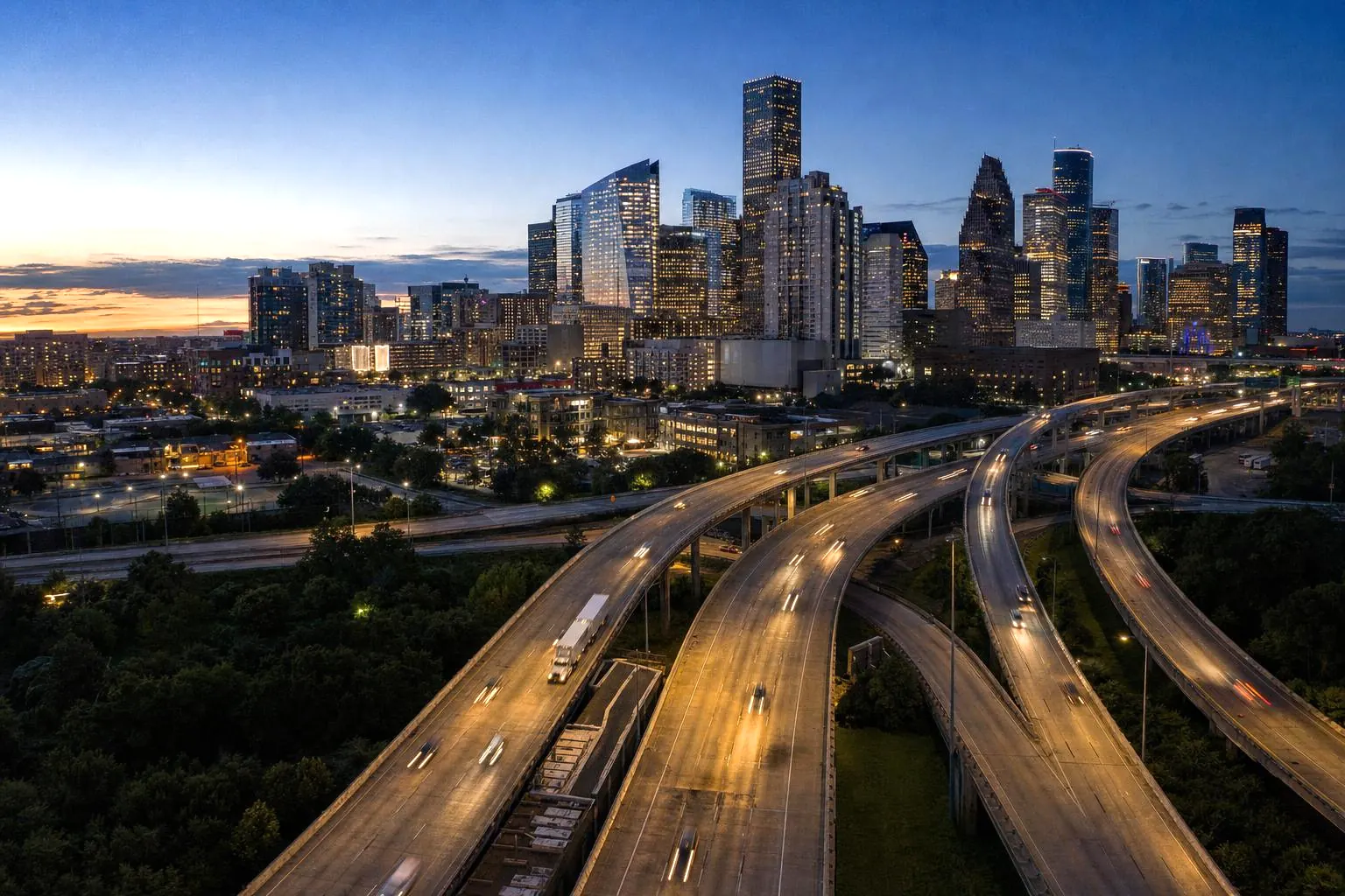 An elevated highway interchange flowing into a downtown skyline at blue hour, with trucks and cars tracing light trails
